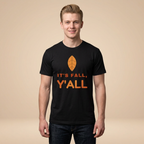 A man wearing a black crewneck t-shirt with an orange graphic that reads 'IT'S FALL, Y'ALL' beneath an orange leaf icon, set against a solid beige background.