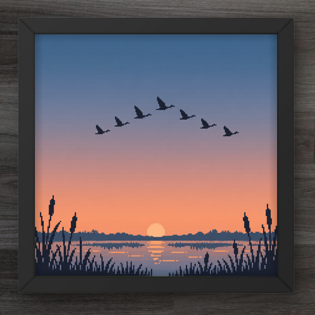 A framed square art print depicting a sunset landscape with a flock of birds flying over a lake, surrounded by reeds, shown on a dark wood background.
