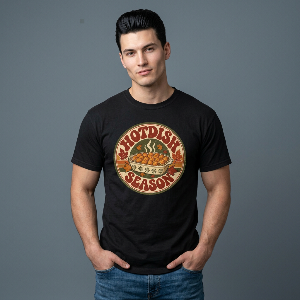 A man wearing a black short-sleeve graphic t-shirt with a circular retro design that reads 'HOTDISH SEASON' around a casserole dish paired with blue jeans, against a solid gray background.
