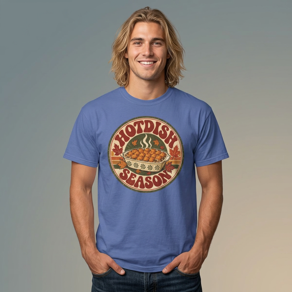 A smiling man with long blonde hair wearing a blue crewneck t-shirt with a circular retro graphic that reads 'HOTDISH SEASON' around a casserole dishl. He is standing against a neutral gray-to-beige gradient background.