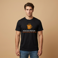 A man wearing a black short-sleeve t-shirt with a coffee-themed graphic that reads 'HOCUS POCUS I NEED COFFEE TO FOCUS' in white and orange text, paired with blue jeans, set against a solid beige background.
