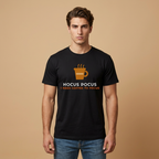 A man wearing a black short-sleeve t-shirt with a coffee-themed graphic that reads 'HOCUS POCUS I NEED COFFEE TO FOCUS' in white and orange text, paired with blue jeans, set against a solid beige background.