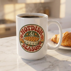 A white ceramic coffee mug with a 'Hotdish Season' graphic, filled with black coffee and sitting on a marble countertop next to a croissant.