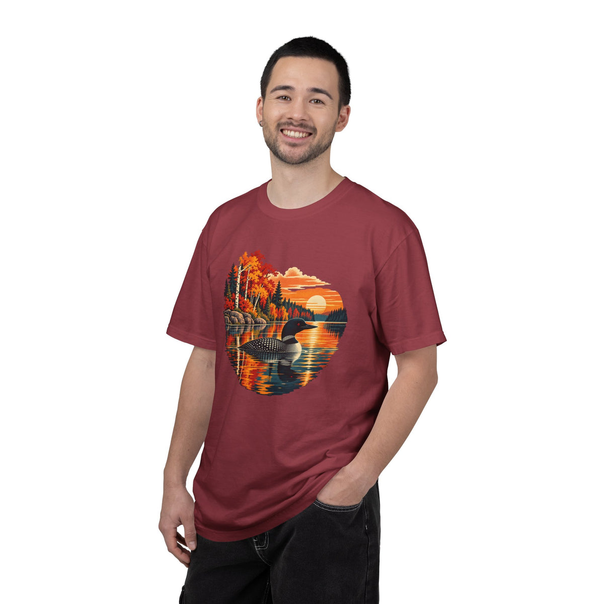 A man wearing a maroon crewneck t-shirt with a circular graphic print of an autumn landscape with a lake, trees, and a sunset. The background is solid white.