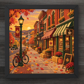 A framed square art print depicting a cozy autumn street scene with brick storefronts, red maple leaf signs, fall leaves, a bicycle, and warm sunset light, shown against a dark wood background.