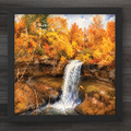 A framed square art print depicting a waterfall flowing into a rocky pool, surrounded by dense autumn trees in bright orange, yellow, and brown tones, set against a dark wood background.