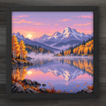 A framed pixel art print of a mountain landscape at sunset, featuring snow-capped peaks, a calm lake with reflections, yellow autumn trees, and a purple sky, displayed on a dark wood background.