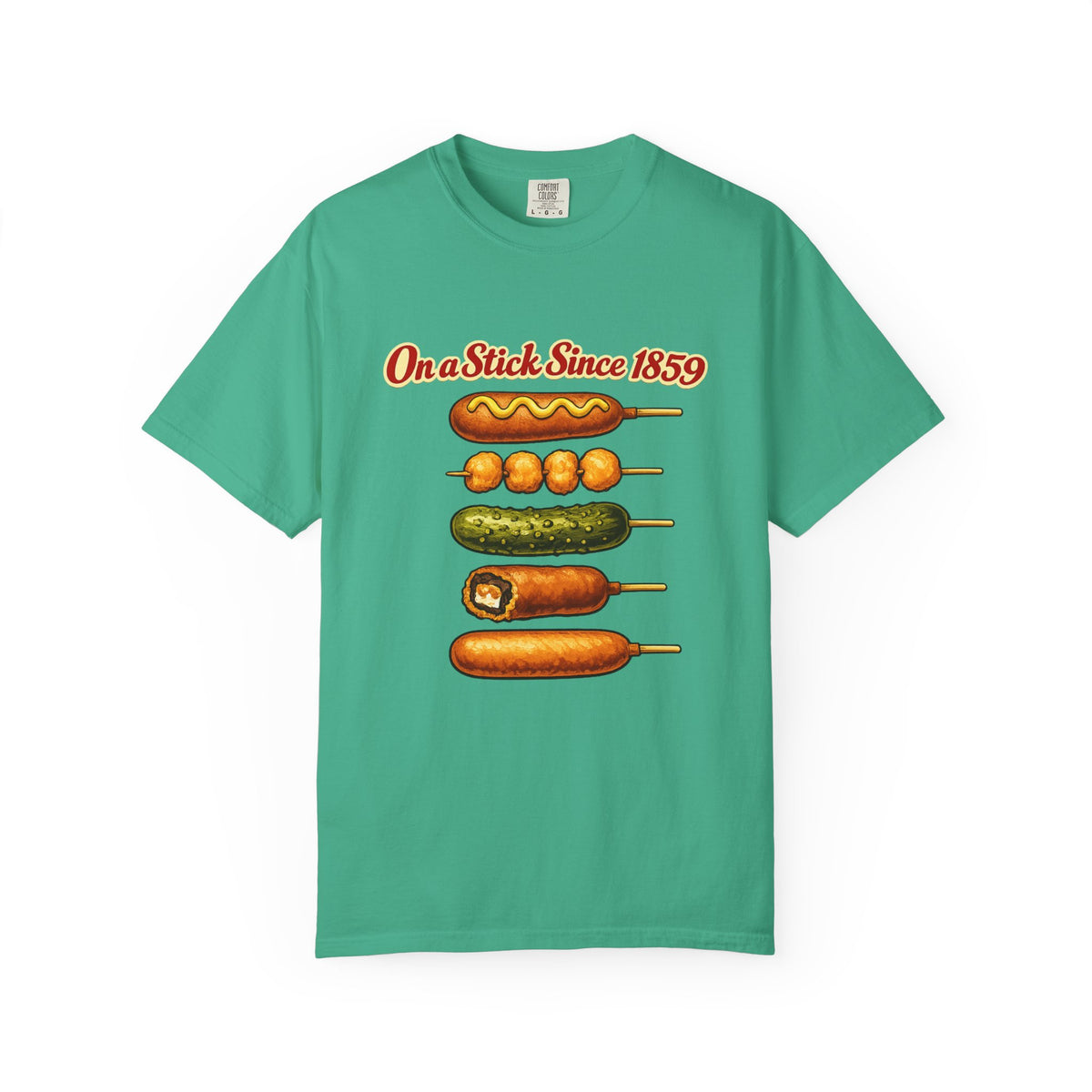 A green short-sleeve crewneck t-shirt featuring a graphic print of various corn dogs and hot dogs stacked vertically with the text 'On a Stick Since 1859' in red above them, displayed on a clean white background.