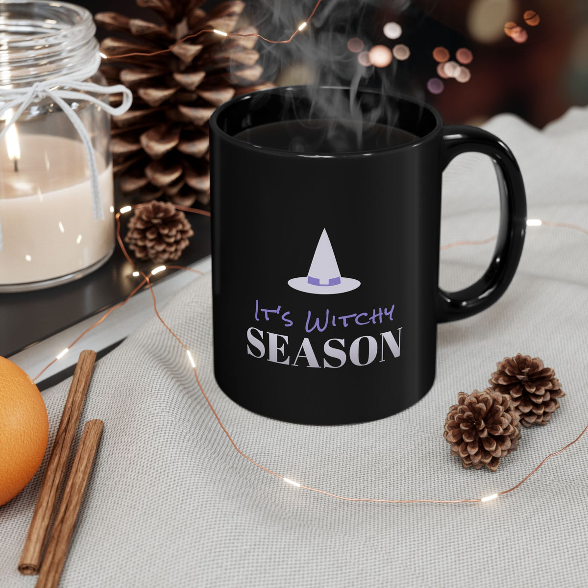 A black mug that says "It's Witchy Season" on a counter next to a candle and pinecones