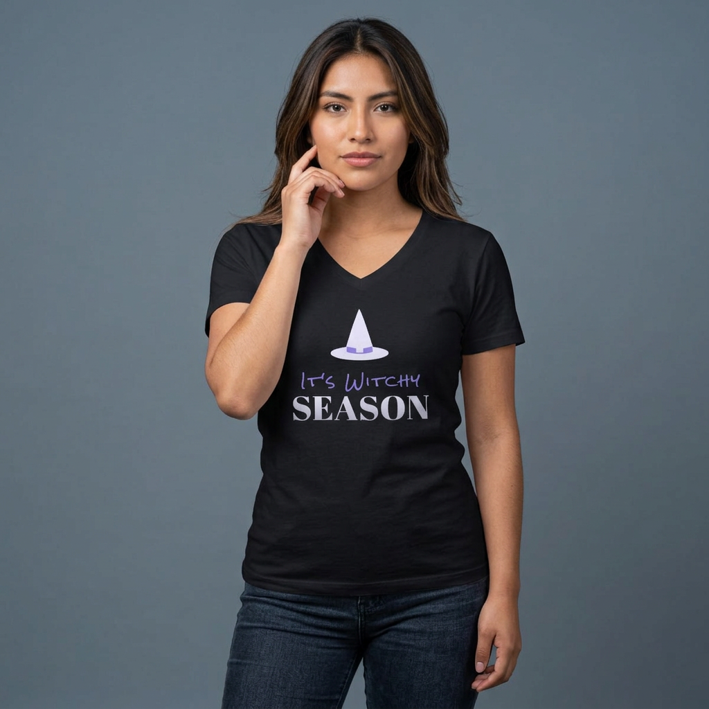 A woman modeling a black V-neck t-shirt with a graphic that reads 'It's Witchy Season' in white and purple text, featuring a small white witch hat illustration above. She is wearing dark blue jeans against a solid gray studio background.